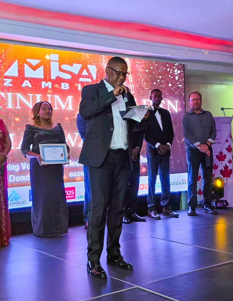 An award presenter speaks on stage beside recipients holding certificates